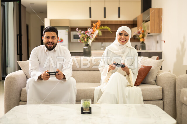 لعب العاب الفيديو الترفيهية، استخدام التكنولوجيا الحديثة، زوجان عربيان سعوديان خليجيان  يجلسان على كنبه مريحه في غرفه المعيشه يمسكان عصا التحكم ، إيماءات التحدي والمتعة و التسليه .