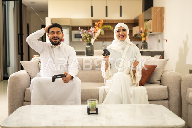 الاستمتاع والتسلية في العطلة ، استخدام التكنولوجيا الحديثة، زوجان عربيان سعوديان خليجيان  يجلسان على كنبه مريحه في غرفه المعيشه يمسكان عصا التحكم ، إيماءات الفوز والخسارة فى اللعبة .