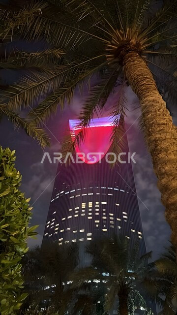 Kingdom Tower lit up in red in Riyadh at night, development and urban progress of facilities in the Kingdom of Saudi Arabia, interest in planting palm trees and green plants in tourist cities, engineering art of towers and skyscrapers in a modern and advanced style