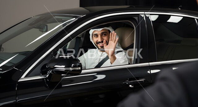 رفع كف اليد بتعابير القاء التحية والسلام، وسائل النقل والمواصلات الحديثة، التنقل باستخدام السيارة الخاصة، رجل عربي خليجي اماراتي مبتسم يرتدي الغترة والكندورة يجلس داخل سيارته، ايماءات السعادة والسرور