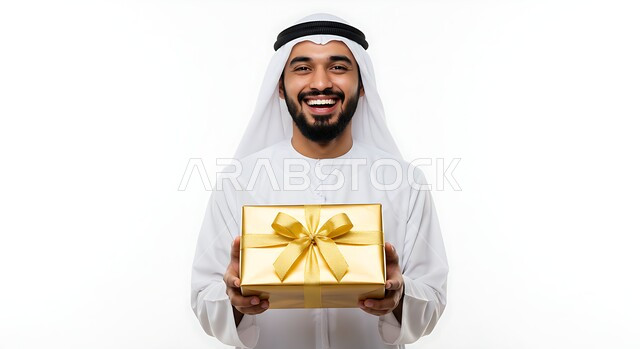 النظر الى الكاميرا بتعابير السرور والفرح، الاحتفال وتقديم الهدايا، الأعياد والمناسبات السعيدة، بورتريه مقرب لرجل عربي خليجي اماراتي مبتسم يرتدي الكندورة والغترة يحمل بيده علبة هدية مغلفة، خلفية بيضاء
