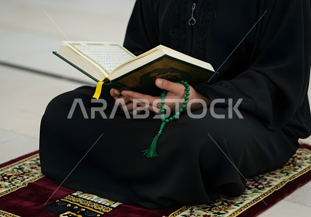 التقرب الى رب العالمين في شهر رمضان المبارك، قراءة كتاب الله تعالى, إيماءات الخشوع و الإجتهاد في العباده, بورتريه مقرب لأيدي امرأة عربية سعودية خليجية ترتدي العباءة السوداء تمسك بيدها السبحة والقرآن الكريم تجلس على سجادة الصلاه, خلفية رمادية