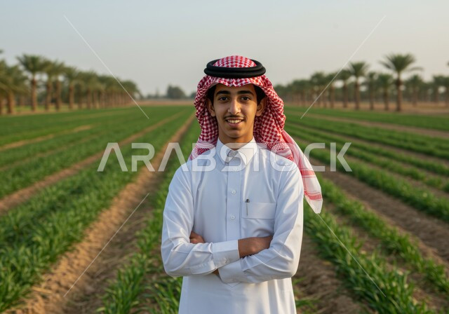 مفهوم الاكتفاء الذاتي، صورة مقربة لرجل عربي خليجي سعودي مبتسم يرتدي الشماغ والثوب التقليدي يقف بأيدي متقاطعة بايماءات الثقة بالنفس والرجولة، النظر الى الكاميرا بتعابير السرور والسعادة، حقل زراعي اخضر في النهار، خلفية الطبيعة