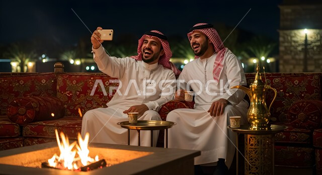 كشتة شعبية شبابية، توثيق اللحظات السعيدة، مفهوم حسن الضيافه والاستقبال، رجل عربي خليجي سعودي يقوم بالتقاط صور السيلفي التذكارية مع صديقه باستخدام الهاتف الجوال، صديقان عربيان خليجيان سعوديان في جلسة بر وتخييم خارجية في الأجواء الشتوية