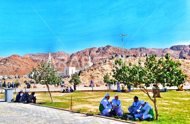 استراحة الحجاج والمعتمرين على العشب امام جبل احد في المدينه المنوره في النهار، القمم والمرتفعات الجبلية في المملكة, جذب و استقطاب السكان من كافة انحاء العالم، معلم اسلامي تاريخي قديم، السياحه في الاماكن الدينيه المشهورة في السعوديه