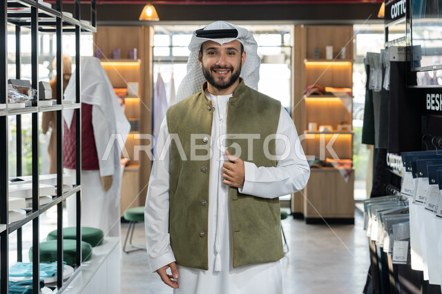 عرض الأزياء والموديلات الحديثة، التبضع وأخذ الحاجات اللازمة، التجول داخل أحد المولات التجارية الفاخرة ، رجل عربى خليجي اماراتي مبتسم يرتدي الكندورة البيضاء يقف مبتسم داخل محل ملابس ، شراء واقتناء المستلزمات من المول