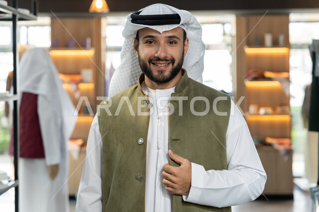شراء واقتناء المستلزمات من المول, التبضع وأخذ الحاجات اللازمة، التجول داخل أحد المولات التجارية الفاخرة ، صورة مقربة لرجل عربى خليجي اماراتي مبتسم يرتدي الكندورة البيضاء يقف مبتسم داخل محل ملابس، عرض الأزياء والموديلات الحديثة