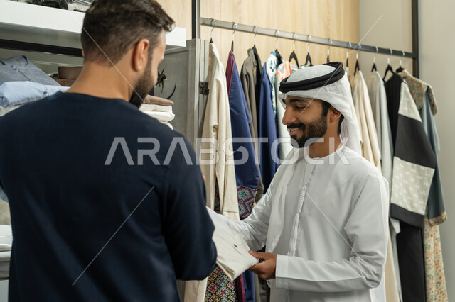 الاستمتاع بقضاء وقت ممتع في التسوق والشراء، اقتناء ملابس جديدة، شاب عربي خليجي اماراتي متبسم يرتدي الكندورة الكحلي يعمل في مهنة الخياطة يقوم بعرض قميص ابيض للزبون، محل خياطة و تفصيل الملابس الرجالي ، مهارة الخياطة واتقان تفصيل الملابس الشبابية