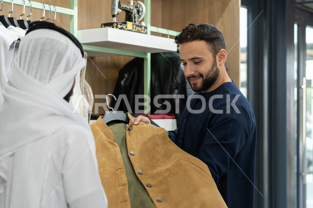 محل خياطة و تفصيل الملابس الرجالي،اختيار أجود أنواع الأقمشة لصنع الثياب، متعه التسوق و شراء الملابس، شاب عربي خليجي اماراتي يرتدي الكندورة الكحلي يعمل في مهنة الخياطة يقوم بعرض جاكيت شتوي للزبون
