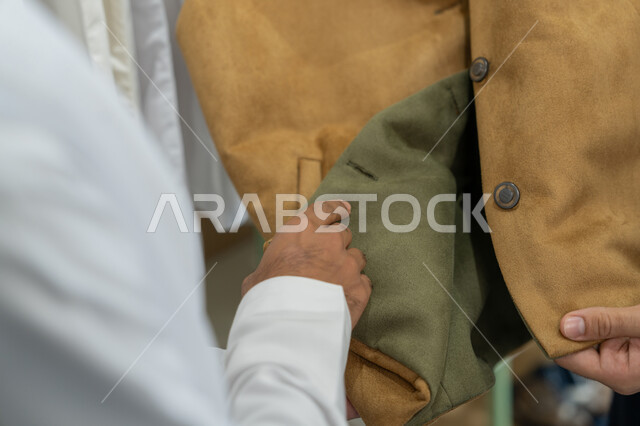 اختيار الأقمشة المناسبة لفصل الشتاء,صورة مقربه ليد رجل عربي خليجي اماراتي يرتدي الكندورة البيضاء يقوم بتفحص جودة خياطه الجاكيت, ورشة عمل تصميم أزياء, مهارة الخياطة واتقان تفصيل الملابس الشبابية
