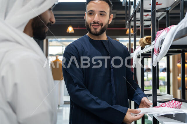 مهارة الخياطة واتقان تفصيل الملابس الشبابية، اختيار أجود أنواع الأقمشة لصنع الثياب، شاب عربي خليجي اماراتي يرتدي الكندورة الكحلي يعمل في مهنة الخياطة يعرض للزبون اجود انواع الغتر ، محل خياطة و تفصيل الملابس الرجالي ،