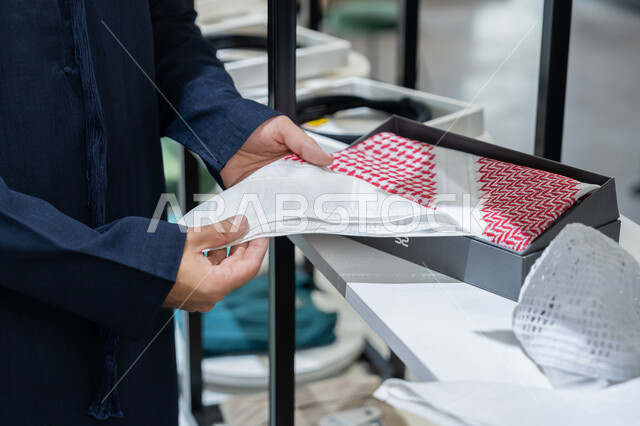 إتقان صنع الملابس، التسوق داخل متجر لوازم الزي الخليجي, صورة مقربة ليد شاب عربي خليجي اماراتي يرتدي الكندورة الكحلي يقوم بعرض  الغتره البيضاء ، محل خياطة و تفصيل الملابس الرجالي، اختيار افضل انواع الاقمشه