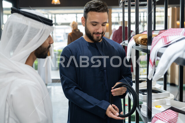 محل خياطة و تفصيل الملابس الرجالي، شاب عربي خليجي اماراتي يعمل في مهنة الخياطة يرتدي الكندورة الكحلي يعرض للزبون اجود انواع الغتره ، مهارة الخياطة واتقان تفصيل الملابس الشباب، الاستمتاع بالتسوق و شراء الاكسسورات
