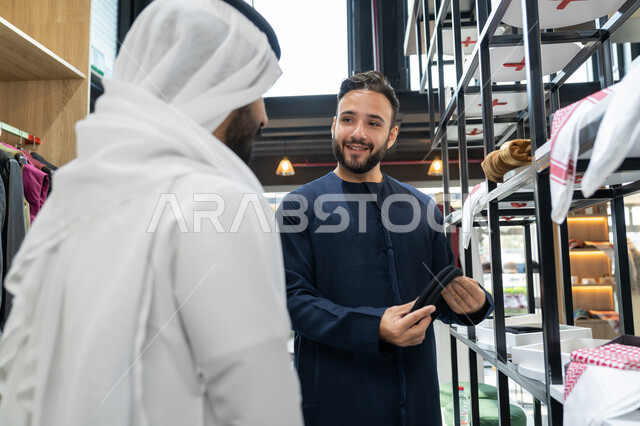 الاستمتاع بالتسوق و شراء الاكسسورات، محل خياطة و تفصيل الملابس الرجالي، شاب عربي خليجي اماراتي يعمل في مهنة الخياطة يرتدي الكندورة الكحلي يعرض للزبون اجود انواع الغتره ، مهارة الخياطة واتقان تفصيل الملابس الشباب