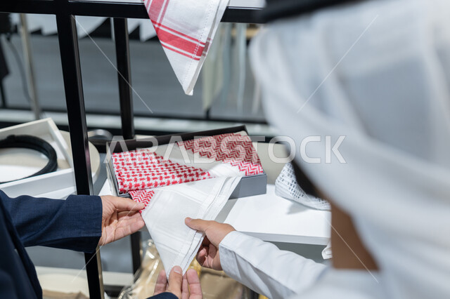 الاستمتاع بالتسوق و شراء الملابس، اتقان تفصيل الملابس، صورة مقربة من الخلف لشاب عربي خليجي اماراتي يرتدي الكندورة الكحلي يعمل في مهنة الخياطة يقوم بعرض للزبون اجود انواع الأقنشة لتفصيل الثوب ، محل خياطة و تفصيل الملابس الرجالي