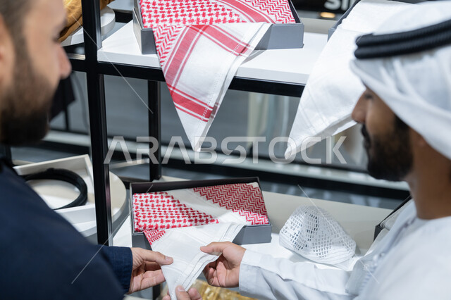 اتقان تفصيل الملابس، صورة مقربة لشاب عربي خليجي اماراتي يرتدي الكندورة الكحلي يعمل في مهنة الخياطة يقوم بعرض اجود انواع الأقمشة لتفصيل الثوب ، محل خياطة و تفصيل الملابس الرجالي