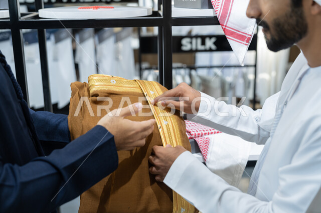 اختيار اجود انواع الأقمشة لتفصيل الثوب، محل خياطة و تفصيل الملابس الرجالي في الامارات، صورة مقربة لشاب عربي خليجي اماراتي يرتدي الكندورة البيضاء داخل محل مختص بخياطة الملابس الرجالية ، مهارة الخياطة واتقان تفصيل الملابس الشبابية