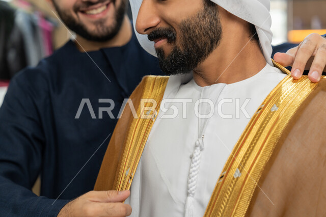 أخذ القياسات المناسبة، محل خياطة و تفصيل الملابس الرجالي ، اختراع وابتكار موديلات جديدة وعصرية، حرف ومهن يدوية، صورة مقربة لشاب خليجي اماراتي يرتدي الكندورة البيضاء يقوم بقياس البشت، فن الخياطة وتصميم ازياء