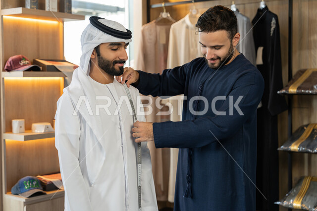 Practicing the tailoring profession, taking appropriate measurements using a tape measure, inventing and creating new and modern models, handicrafts and crafts, a young Emirati man wearing a navy blue kandura taking measurements of the customer's body, a men's tailoring and tailoring shop