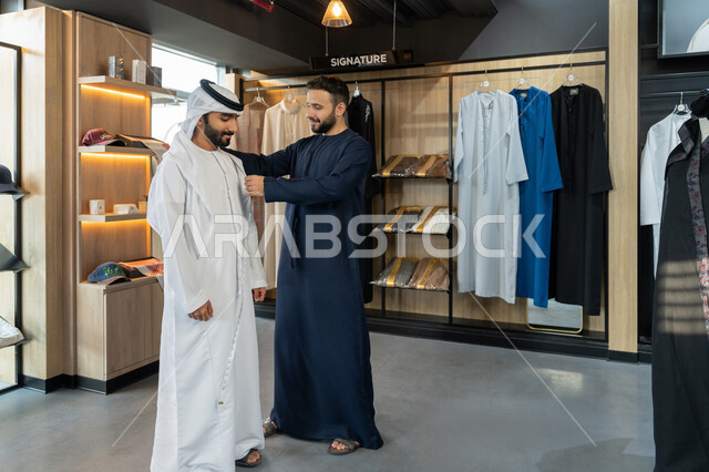 أخذ القياسات المناسبة باستخدام الشريط المتري، اختراع وابتكار موديلات جديدة وعصرية، حرف ومهن يدوية، ممارسة مهنة الخياطة، شاب خليجي اماراتي يرتدي الكندورة الكحلي يقوم بأخذ القياسات الخاصة بجسم الزبون ، محل خياطة و تفصيل الملابس الرجالي ، الجسم بالطول الكامل