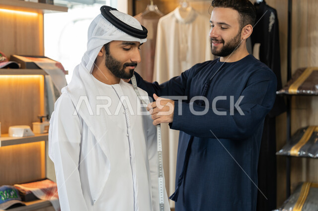اختراع وابتكار موديلات جديدة وعصرية، حرف ومهن يدوية، ممارسة مهنة الخياطة شاب خليجي اماراتي يرتدي الكندورة الكحلي يقوم بأخذ القياسات الخاصة بجسم الزبون  باستخدام الشريط المتري ، محل خياطة و تفصيل الملابس الرجالي