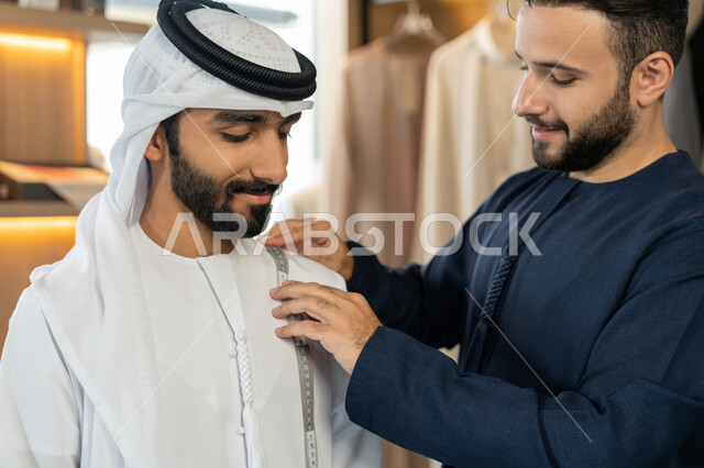 مهارة الخياطة واتقان تفصيل الملابس الشبابية، حرف ومهن يدوية، ممارسة مهنة الخياطة،صورة مقربة لشاب خليجي اماراتي يرتدي الكندورة الكحلي يقوم بأخذ القياسات الخاصة بجسم الزبون  باستخدام الشريط المتري ، محل خياطة و تفصيل الملابس الرجالي