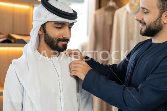 محل خياطة و تفصيل الملابس الرجالي ، حرف ومهن يدوية، ممارسة مهنة الخياطة،صورة مقربة لشاب خليجي اماراتي يرتدي الكندورة الكحلي  يقوم بأخذ القياسات الخاصة بجسم الزبون ر باستخدام الشريط المتري ، مهارة الخياطة واتقان تفصيل الملابس الشبابية