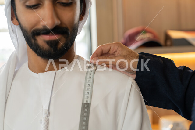 حرف ومهن يدوية، ممارسة مهنة الخياطة، صورة مقربة ليد شاب خليجي اماراتي يرتدي الكندورة الكحلي  يقوم بأخذ القياسات الخاصة بجسم الزبون باستخدام الشريط المتري ، محل خياطة و تفصيل الملابس الرجالي  ، مهارة الخياطة واتقان تفصيل الملابس الشبابية