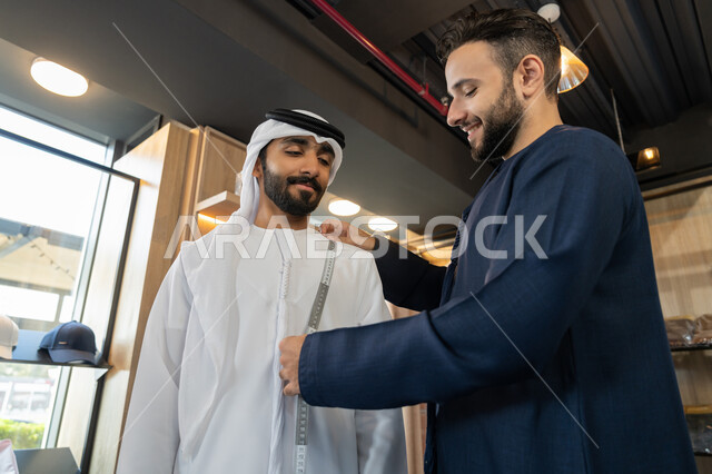 ممارسة مهنة الخياطة، متابعة الموضه ، استعمال أدوات ومعدات الخياطة، حرف ومهن يدوية، خياط عربي خليجي اماراتي يرتدي الكندورة الزرقاء يستخدم شريط القياس لتحديد المقاسات، متعه التسوق في المول