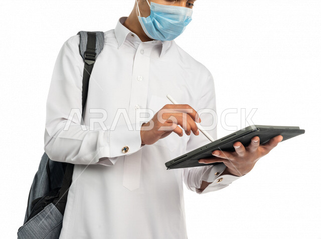 بورتريه لطالب جامعي عربي خليجي سعودي يرتدي الكمامة الطبية للوقاية من فايروس كورونا، يستخدم جهاز التابلت لكتابة وتدوين الملاحظات ، متابعة المحاضرات الجامعية الالكترونية، حل الواجبات الالكترونية ، التعليم الالكتروني ، التعليم الجامعي في السعودية