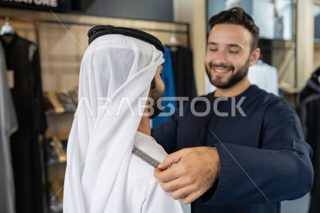 مفهوم الموضة والأناقة، استعمال أدوات ومعدات الخياطة، حرف ومهن يدوية، صورة مقربه لخياط عربي خليجي اماراتي يرتدي الكندورة الزرقاء يقوم باخذ مقاس الزبون، اختيار أجود أنواع الأقمشة لصناعة الأثواب، متعه التسوق في المول