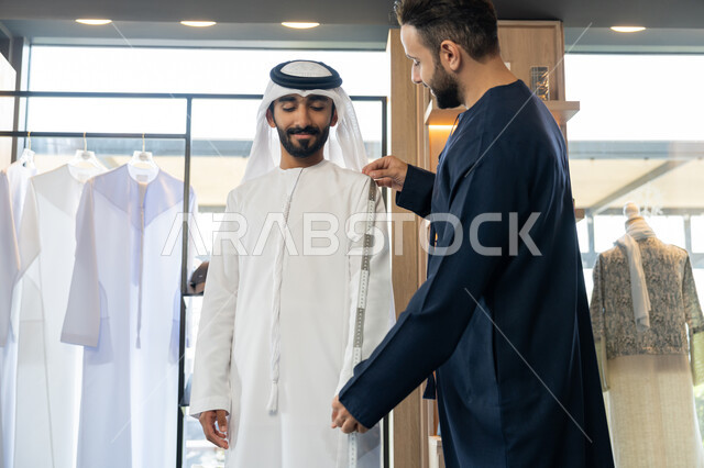 حرف ومهن يدوية، إتقان صنع الملابس ، استعمال أدوات ومعدات الخياطة، صورة مقربه ليد خياط عربي خليجي اماراتي يرتدي الكندورة الزرقاء يقوم باخذ مقاس الزبون، اختيار أجود أنواع الأقمشة لصناعة الأثواب، متعه التسوق في المول