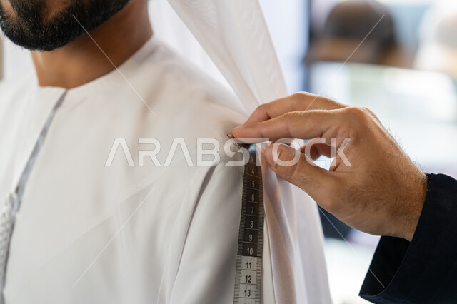 متابعة الموضه ، استعمال أدوات ومعدات الخياطة، حرف ومهن يدوية، صورة مقربه ليد خياط عربي خليجي اماراتي يرتدي الكندورة الزرقاء يقوم باخذ مقاس الزبون، اختيار أجود أنواع الأقمشة لصناعة الأثواب، متعه التسوق في المول
