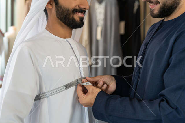 التسوق من الماركات العالميه، استعمال أدوات ومعدات الخياطة، حرف ومهن يدوية، صورة مقربه لخياط عربي خليجي اماراتي يرتدي الكندورة الزرقاء يقوم باخذ مقاس الزبون، اختيار أجود أنواع الأقمشة لصناعة الأثواب، متعه التسوق في المول