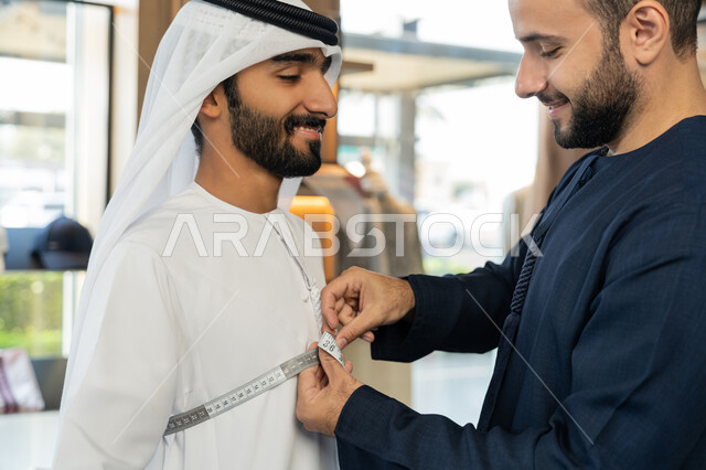 متابعة أحدث صيحات الموضة، استعمال أدوات ومعدات الخياطة، حرف ومهن يدوية، صورة مقربه لخياط عربي خليجي اماراتي يرتدي الكندورة الزرقاء يقوم باخذ مقاس الزبون، اختيار أجود أنواع الأقمشة لصناعة الأثواب، متعه التسوق في المول