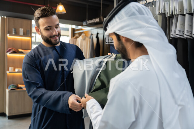 محل خياطة و تفصيل الملابس الرجالي، بائع يعرض للزبون اجود انواع الاقمشة ، مهارة الخياطة واتقان تفصيل الملابس الشبابية، رجل عربى خليجي اماراتي يرتدي الزي التقليدي يقوم بشراء الملابس