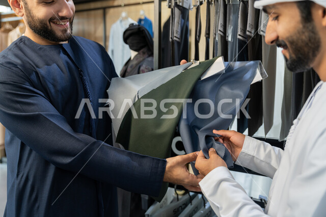 الاستفادة من العروض والتخفيضات الموسميه، تعابير وايماءات السرور والسعادة، عرض الأزياء والموديلات الحديثة،  صورة مقربة لرجل عربي خليجي اماراتي يرتدي الثوب التقليدي يقوم بشراء الملابس، بائع يعرض للزبون اجود انواع الأقمشة لاخيارالثوب، متعه التسوق و شراء الملابس الجديده