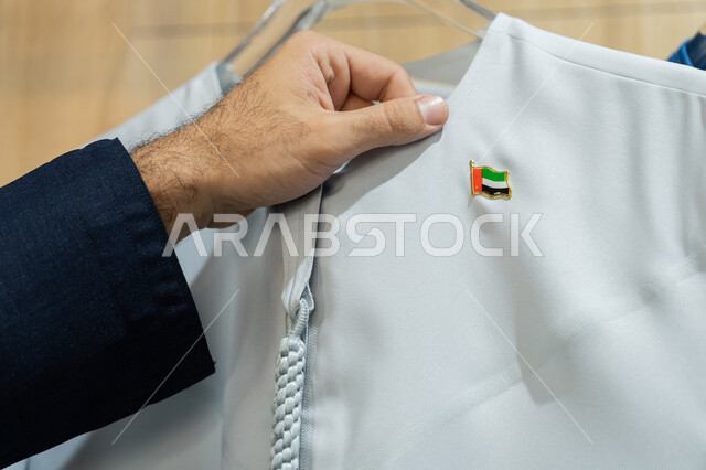 شراء الاحتياجات والمستلزمات الرجالية من المتجر، صورة مقربة ليد رجل عربي خليجي اماراتي يرتدي الزي التقليدي يقوم بالتسوق في المول ،شاب يقوم بلمس كندورة بيضاء عليها علم الامارات المتحده العربية، عرض الأزياء والموديلات الحديثة