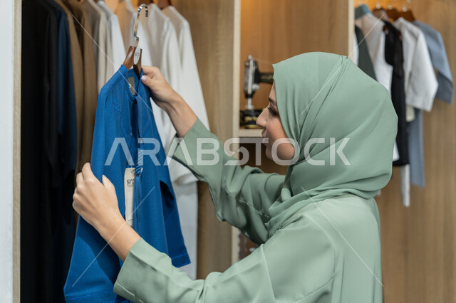 شراء الاحتياجات والملابس, التسوق داخل مول تجاري , صورة من الجانب لامراة عربيه خليجيه اماراتية ترتدي العباءة الملونه تقوم بأختيار ملابس، متابعة الموضه، سيده سعوديه تقوم باختيار عباءه باللون الازرق , فن تصاميم العباءات العصرية الخليجية المميزة