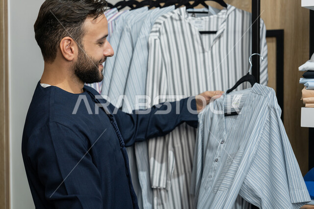 الاستفادة من العرض و الخصم الموسمي,  صورة مقربة لزبون عربي خليجي اماراتي مبتسم يرتدي كندورة كحلي يقوم باختيار ثوب من متجر من متجر المستلزمات الرجاليه , إيماءات السعادة و السرور، الاستمتاع بالتسوق في المول التجاري في الامارات العربيه المتحده