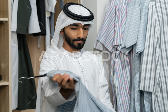 قضاء وقت ممتع في شراء الملابس الرجالية، رجل عربي خليجي اماراتي يرتدي كندوره بيضاء و غتره داخل المتجر يقوم باختيار قميص، الاستمتاع بالتسوق والاستفاده من العرض و الخصم الموسمي