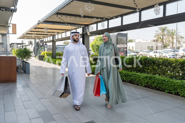 الاستفادة من العرض الخصم في المول، التعاون والتشاور بين الزوجين, زوجان عربيان خليجان اماراتيان يرتديان الزي التقليدي يقومان بالتسوق وحمل اكياس التسوق، متعه التسوق و شراء الملابس الجديده، صورة الجسم بالطول الكامل