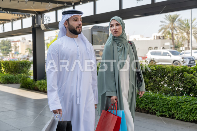 Cooperation and consultation between spouses, purchasing supplies from the mall, taking advantage of the seasonal discount offer, an Arab Gulf Emirati couple wearing traditional dress shopping and carrying shopping bags, full body image