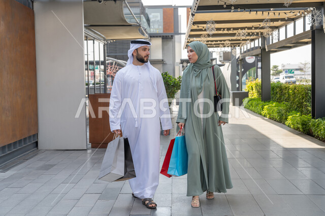 إقتناء المستلزمات من المول, الاستفادة من الخصم و العرض الموسمي, زوجان عربيان خليجيان اماراتيان يرتديان الزي التقليدي يقومان بالتسوق وحمل اكياس التسوق, التعاون والتشاور بين الزوجين، صورة الجسم بالطول الكامل