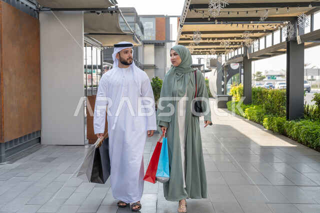 متعة الشراء في الامارات العربية المتحده, الاستفادة من االخصم و العرض الموسمي, زوجان عربيان خليجان اماراتيان يرتديان الزي التقليدي يقومان بحمل اكياس التسوق من المولات والمحلات التجارية الخليجية, التعاون والتشاور بين الزوجين، صورة الجسم بالطول الكامل