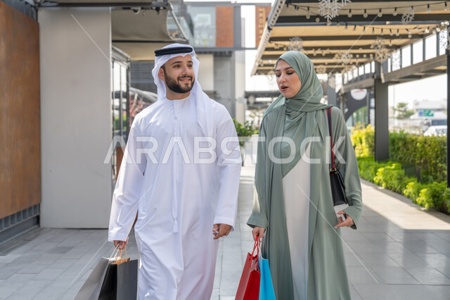 Taking advantage of the offer and discount, cooperation and consultation between spouses, an Arab Gulf Emirati couple wearing traditional dress carrying shopping bags, purchasing supplies from malls, full body image