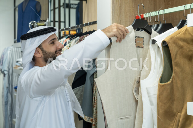 قضاء وقت ممتع في شراء المستلزمات الرجاليه من المتجر، الاستفادة من العرض والخصم، رجل عربي خليجي اماراتي يرتدي كندوره بيضاء و غتره داخل احدى مراكز المول التجاري يقوم باختيار الملابس، الاستمتاع بالشراء والتسوق