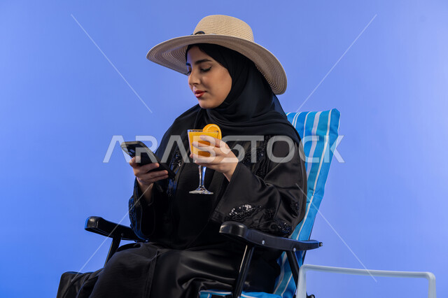 رحلات ترفيهية في المنتجعات البحرية, تناول المشروبات الصيفية المنعشة , بورترية لامراة عربيه خليجيه امارتيه محجبة ترتدي العباءه السوداء و قبعه من القش, امراة سعودية مبتسمة تقوم بالنظر فى الهاتف المحمول, خلفية زرقاء