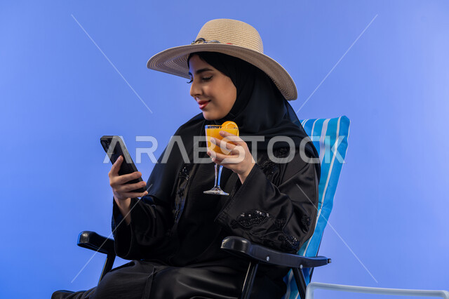 الترفيه والتسلية فى العطلة، الاستمتاع بالمشروبات الطازجة, بورترية لامراة عربيه خليجيه امارتيه محجبة  ترتدي العباءه السوداء و قبعه من القش, امراة سعودية مبتسمة تقوم بالنظر فى الهاتف المحمول , خلفية زرقاء