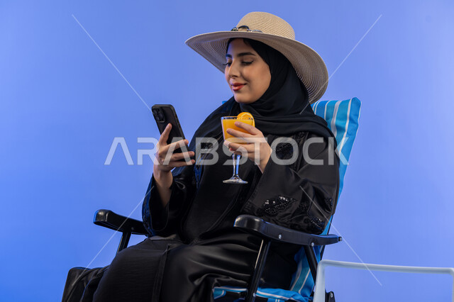 تناول المشروبات الصيفية المنعشة , بورترية لامراة عربيه خليجيه امارتيه محجبة ترتدي العباءه السوداء و قبعه من القش, امراة سعودية مبتسمة تقوم بالنظر فى الهاتف المحمول بجانب حافظة التبريد , خلفية زرقاء